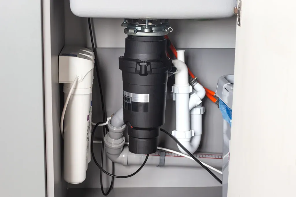 Black garbage disposal mounted under a kitchen sink with white PVC drain piping