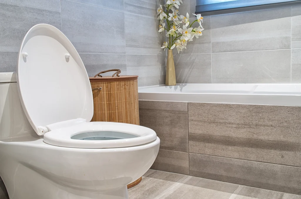 White toilet with the lid raised in a gray tiled bathroom next to a bathtub