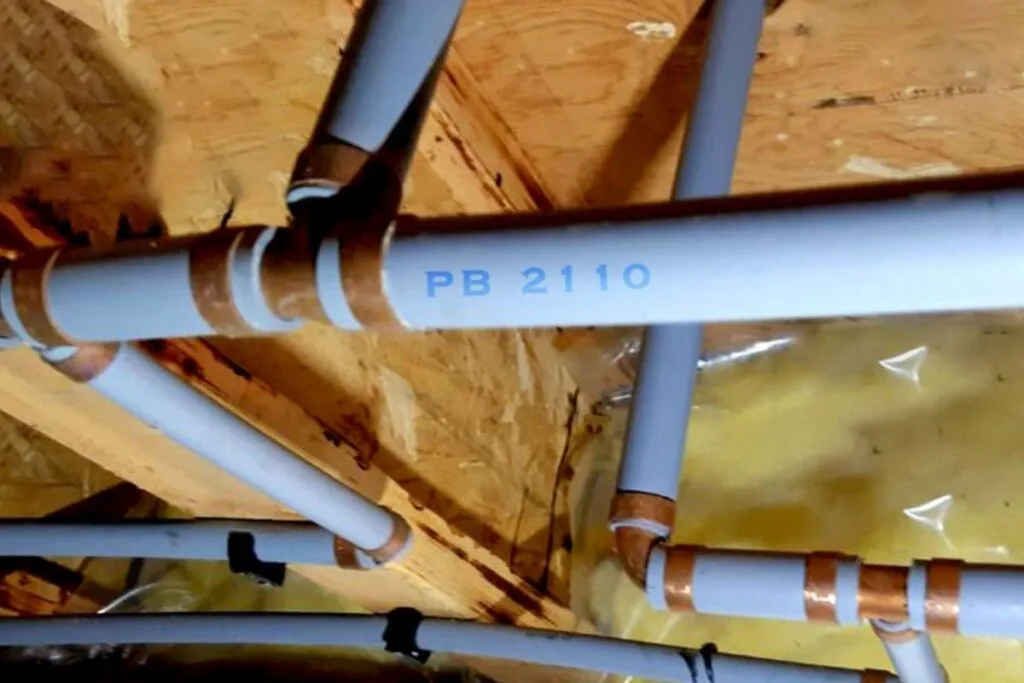 Gray polybutylene supply pipes stamped PB 2110 with copper crimp rings, run along the floor joists