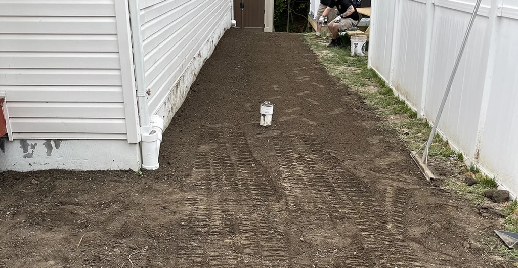 Sewer cleanout pipe capped at ground level in a freshly graded side yard between a house and a vinyl fence