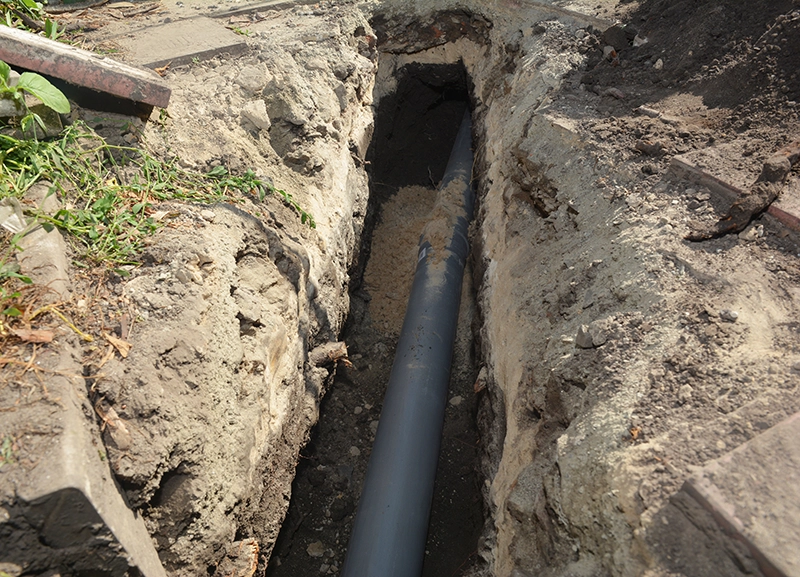 Gray sewer pipe exposed at the bottom of a narrow dug trench