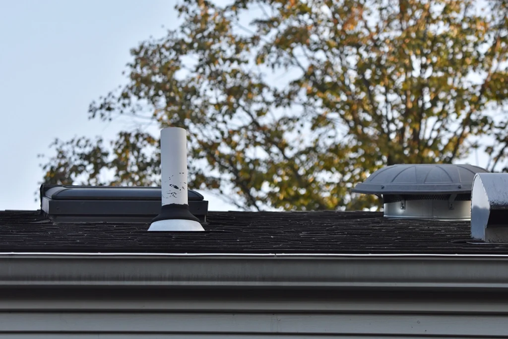 White plumbing vent pipe rising through a shingled roof beside two roof vents