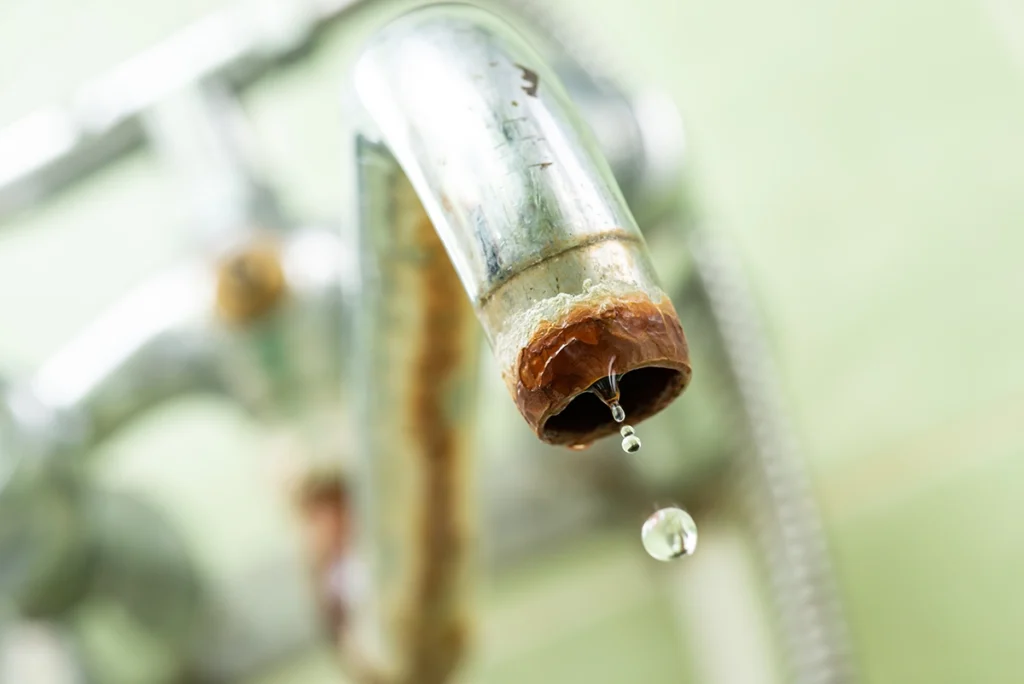 Close-up of a chrome faucet spout crusted with brown corrosion and mineral scale, a drop of water falling from it