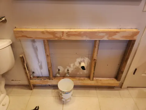 Before: Bathroom wall opened behind a removed vanity, showing the old sink supply stubs and drain fitting