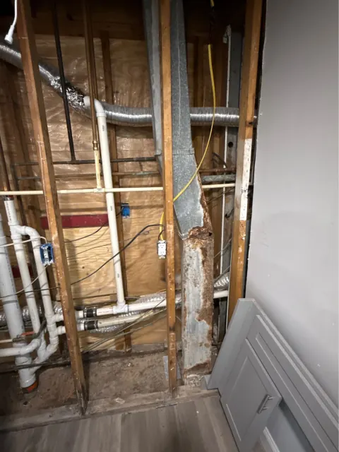 Before: Open wall framing with white drain and vent pipes beside a rusted metal duct