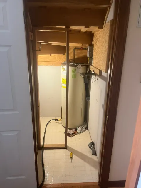 Before: Older tan water heater in an open framed closet with a garden hose on the tile floor