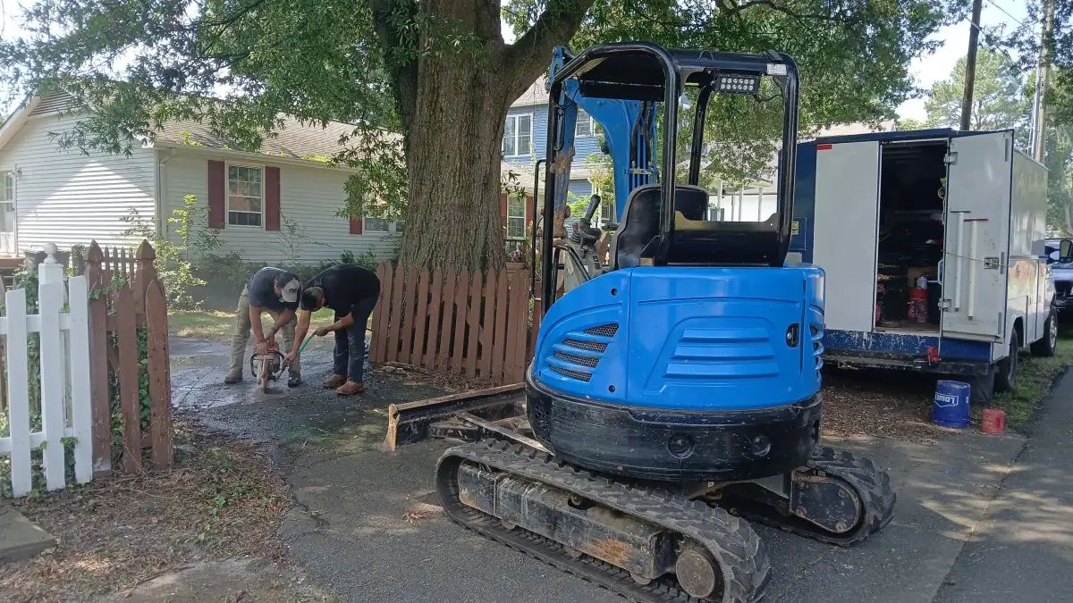 Two-man crew cutting sewer pipe beside a mini-excavator, service van behind.